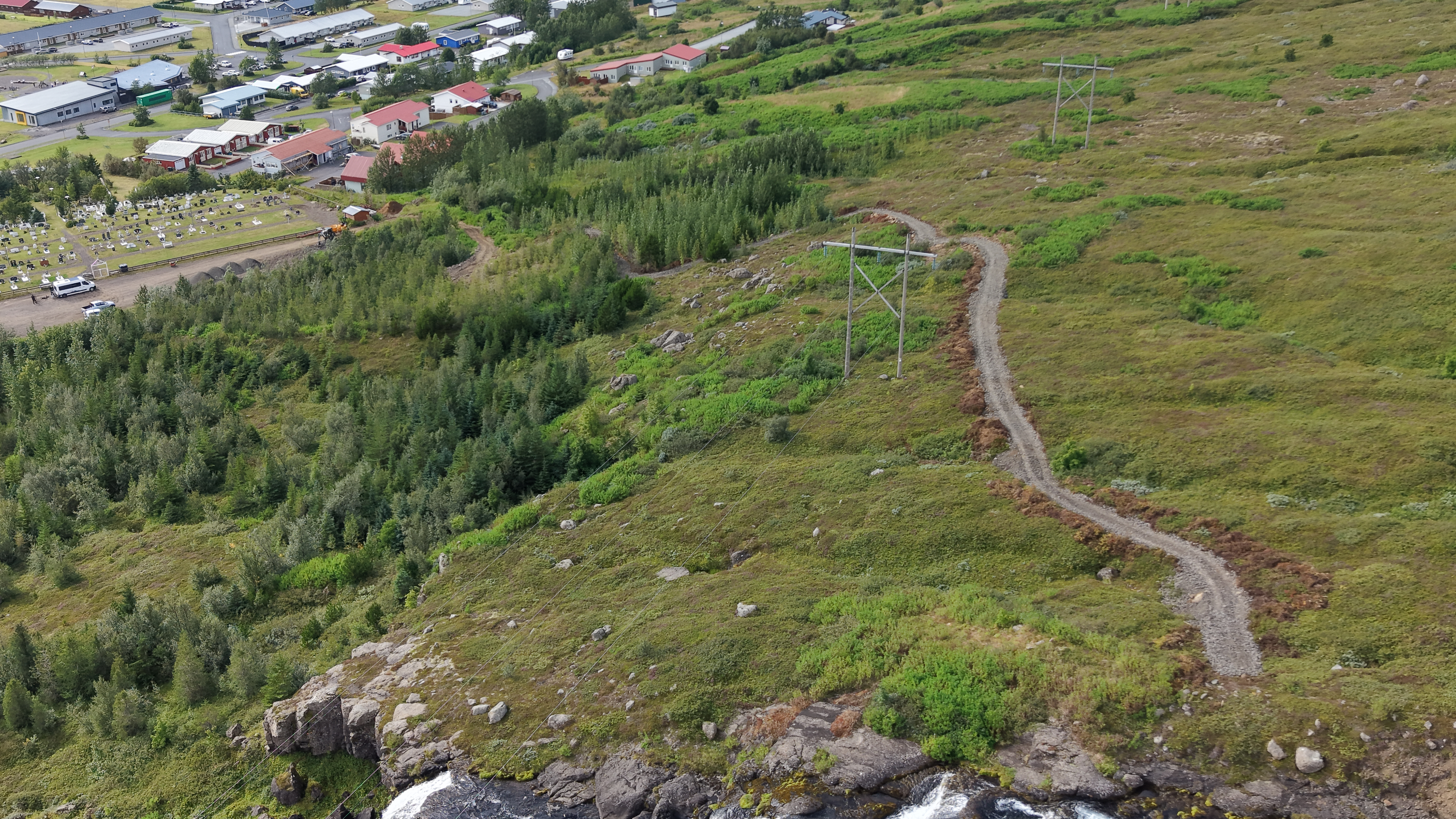 Nýr malarstígur liggur í sveigjum upp gróna hlíð frá Bleiksá. Rafmagnsmöstur standa meðfram stígnum og hluti byggðarinnar á Eskifirði sést í baksýn.